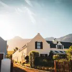 A picturesque row of charming houses with gabled roofs and lush gardens, set against a backdrop of mountains under a clear sky with the sun casting a warm glow.