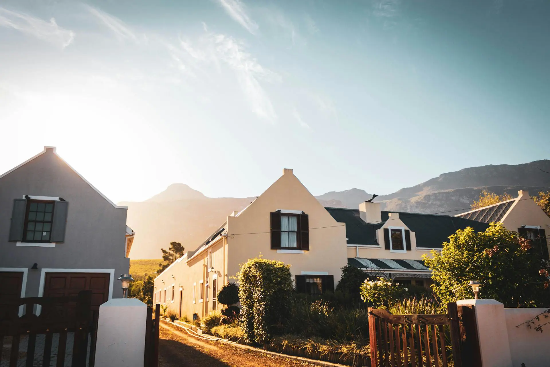 A picturesque row of charming houses with gabled roofs and lush gardens, set against a backdrop of mountains under a clear sky with the sun casting a warm glow.
