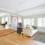 A bright, modern living room with wooden flooring, a cozy sofa, a small table, a desk with a chair, large windows with blinds, and an adjacent room visible through glass doors