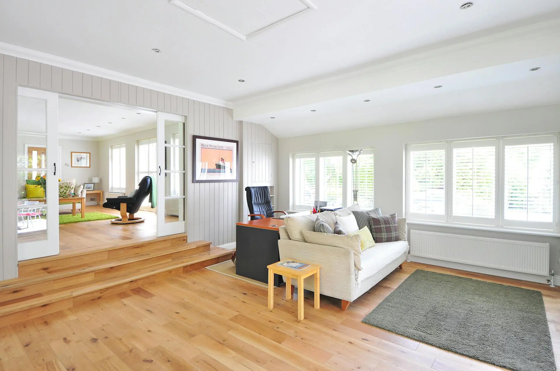 A bright, modern living room with wooden flooring, a cozy sofa, a small table, a desk with a chair, large windows with blinds, and an adjacent room visible through glass doors