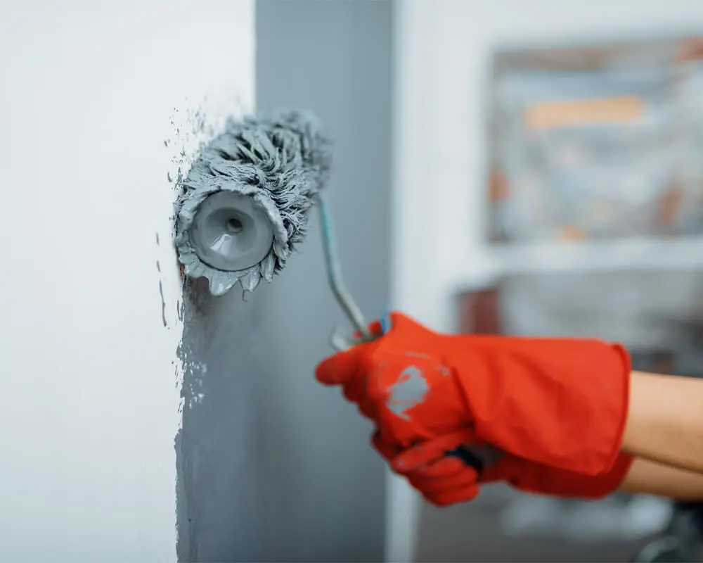 home-about-salute-to-painting A person wearing red gloves paints a wall with a gray paint roller in a room with a blurred background.