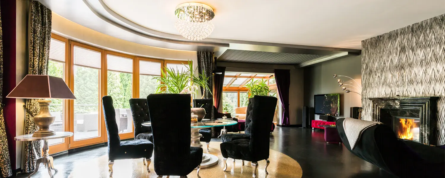 residential-and-commercial-services A luxurious living room with black velvet chairs, a crystal chandelier, large windows, a fireplace with a roaring fire, and elegant decor including plants and a TV.