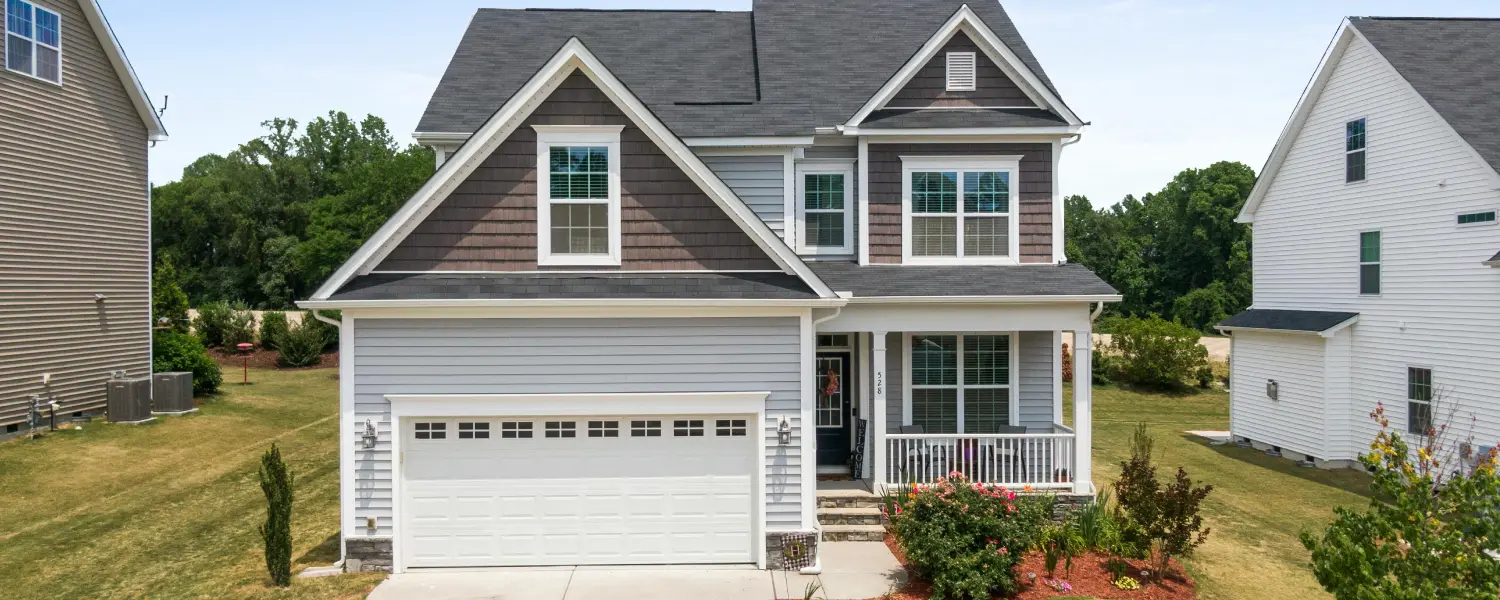 A modern two-story house with a gray exterior, white trim, a double garage, a front porch with columns, and a well-maintained lawn, surrounded by neighboring homes and greenery.