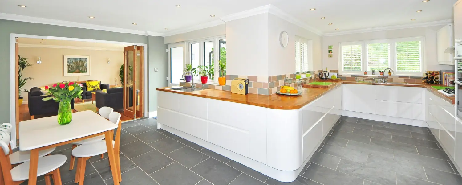 A modern open-plan kitchen with white cabinets, wooden countertops, a dining table with chairs, and an adjacent living room with a sofa and flowers, featuring large windows and tiled flooring.