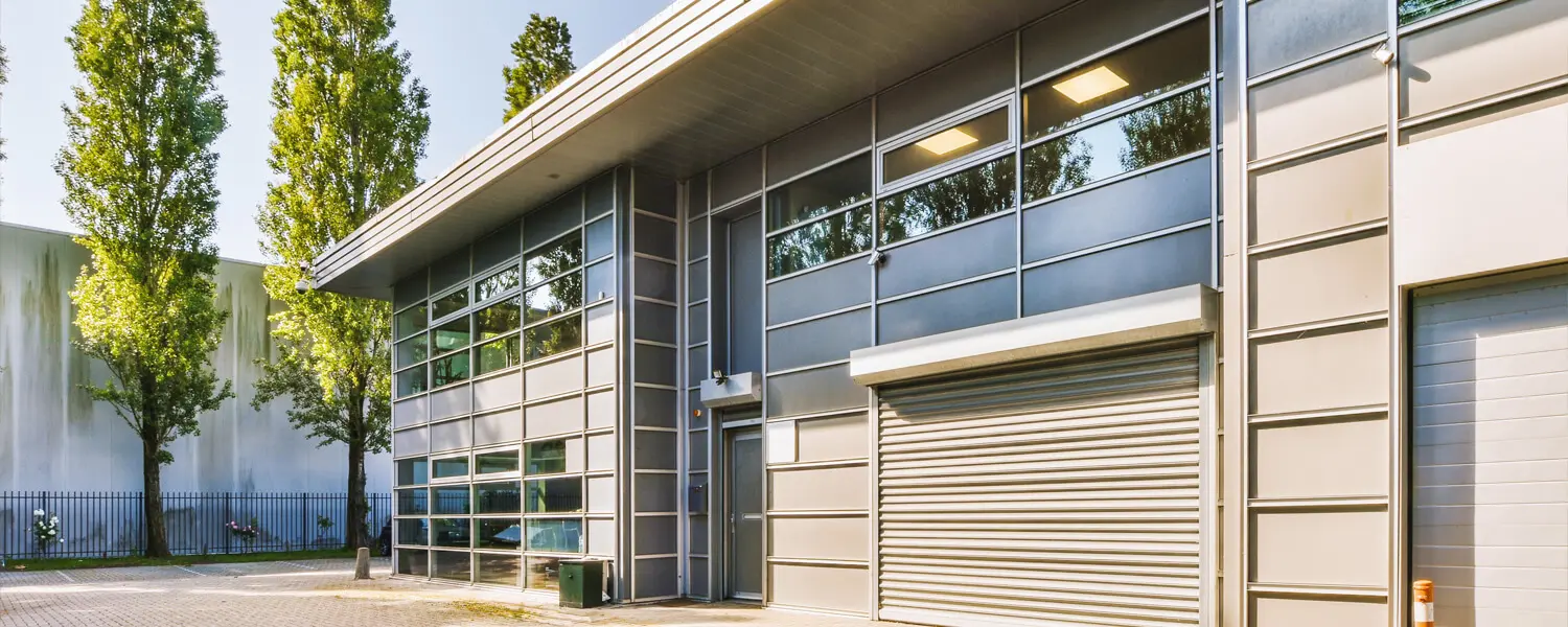 A modern industrial building with large glass windows, a roller shutter door, and a beige and gray facade, surrounded by tall trees and a paved area.