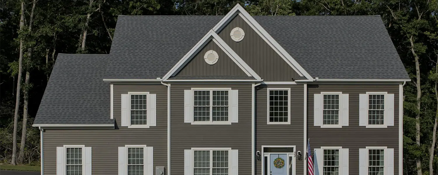A two-story gray house with white trim, multiple windows with shutters, a wreath on the door, an American flag, and a backdrop of tall trees.